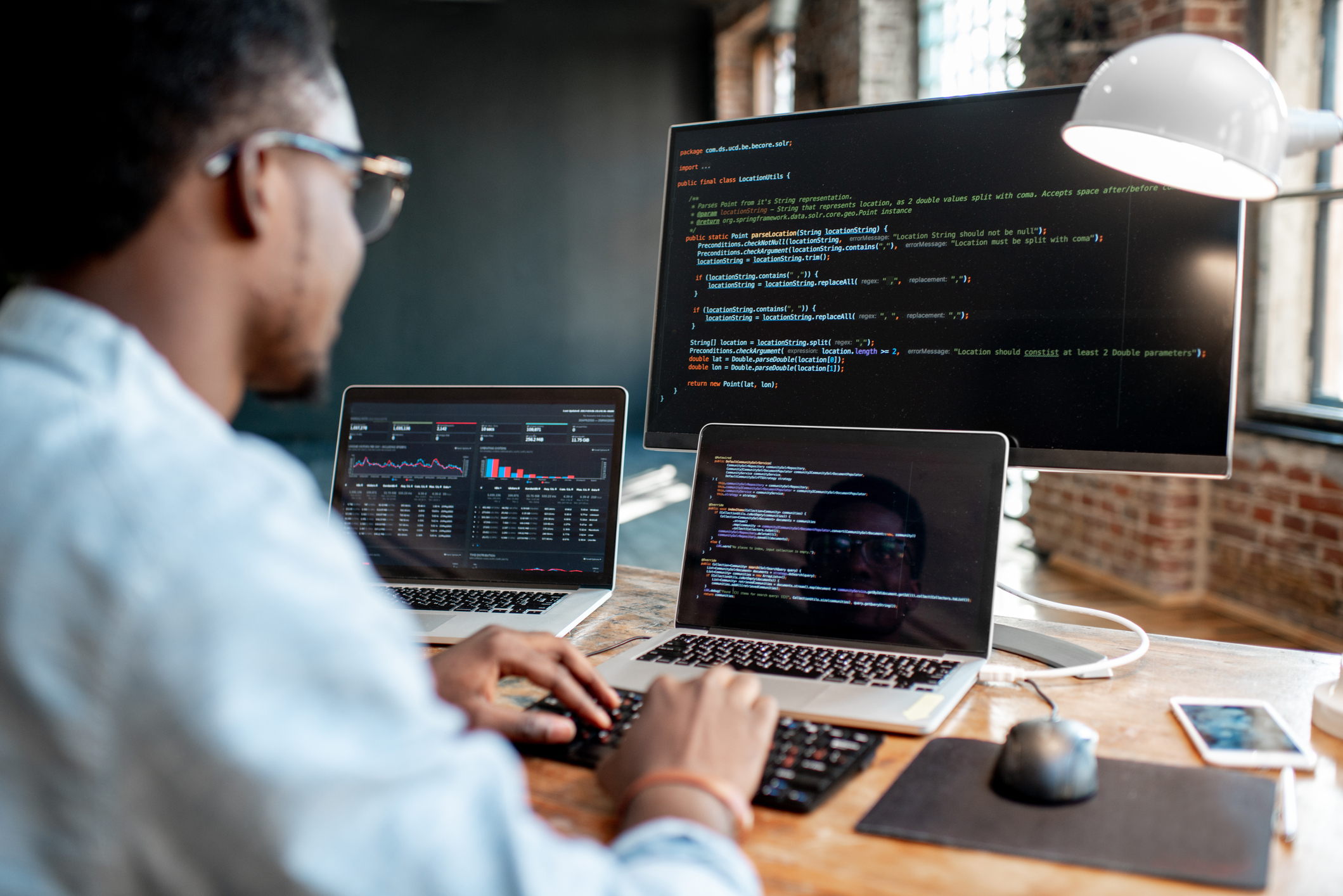 Young african male programmer writing program code sitting at the workplace with three monitors in the office. Image focused on the screen